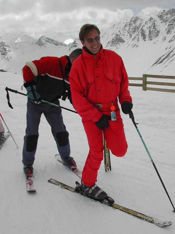 Two people on skis, one in red adjusting gear while the other in black and red helps.