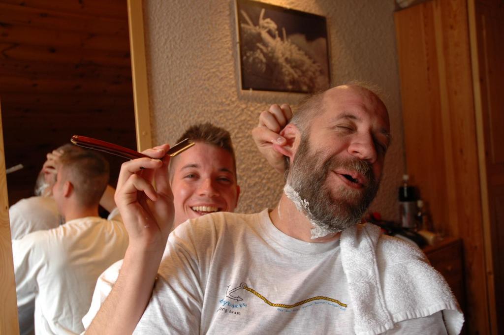 A young man playfully pretends to shave an older man's beard with a straight razor while both smile.