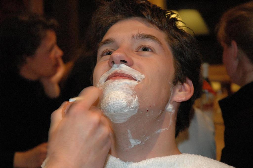 A person applies shaving cream to a young man's face as he tilts his head back.