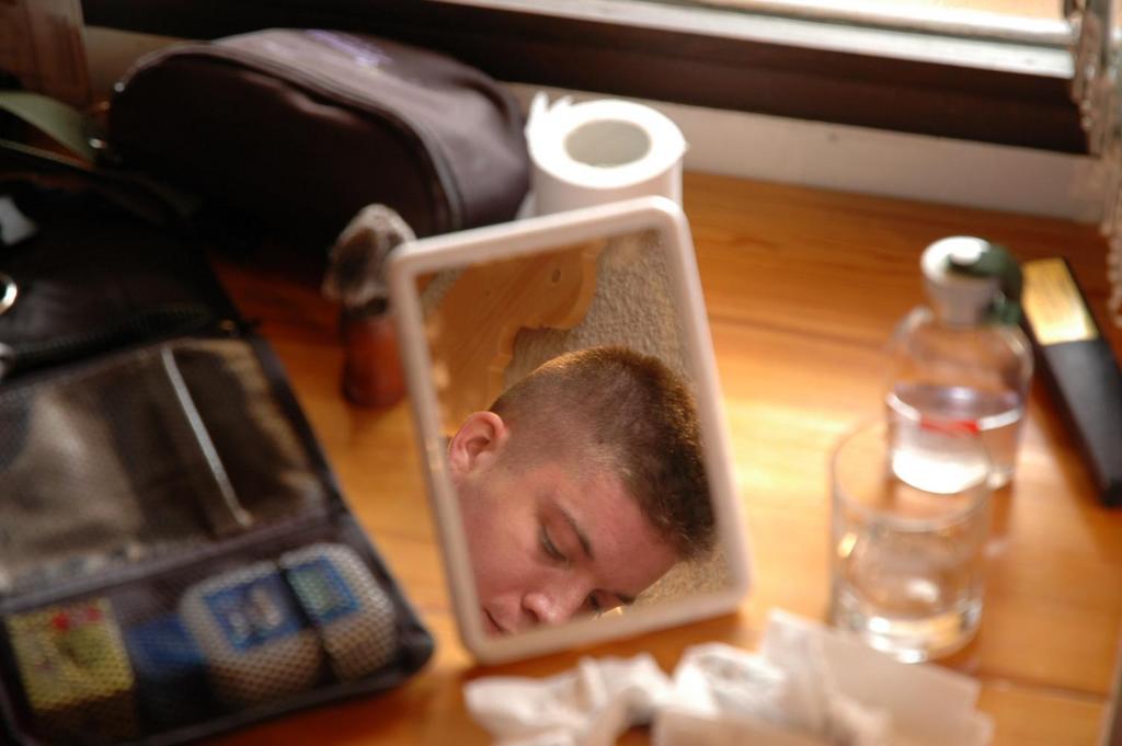 A person's face is reflected in a small mirror on a wooden table with personal items around.