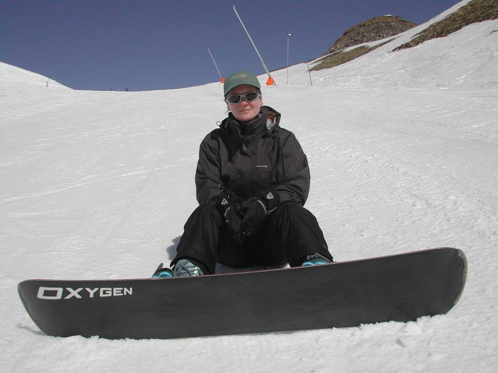 A person in winter gear sits on the snow with a snowboard, wearing sunglasses and a cap.