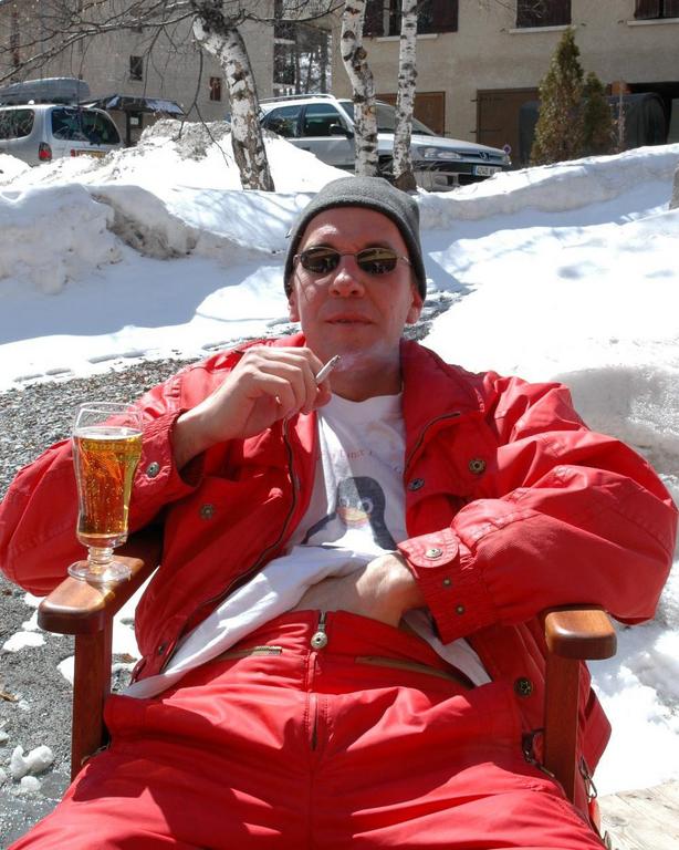 A person in a red jacket relaxes outdoors in the snow, smoking and drinking from a glass.