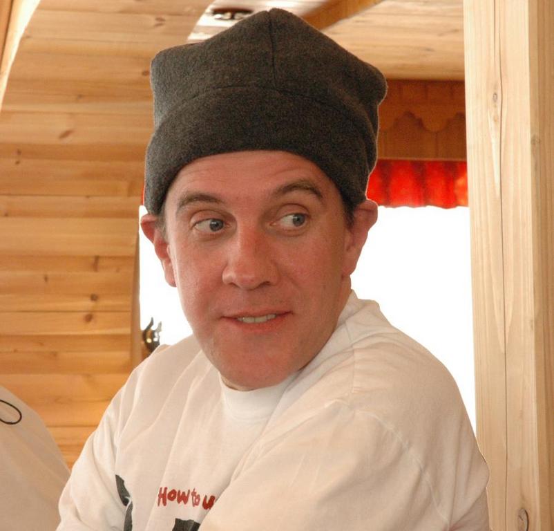 A man wearing a gray hat and white shirt looks to the side with a slight smile indoors.