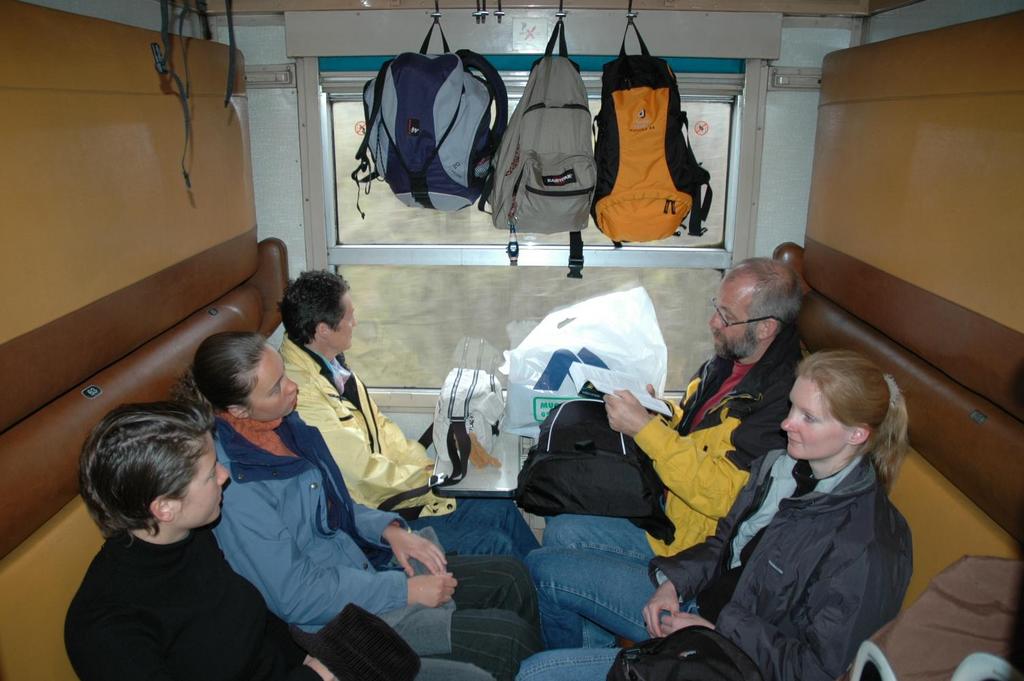 Five people sit in a train compartment, with backpacks hanging above.