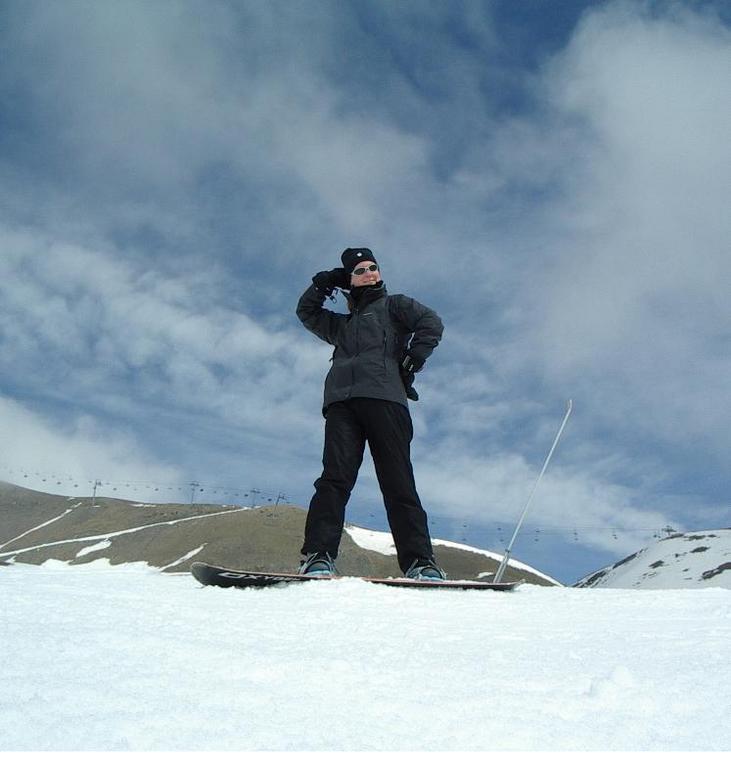 A person in winter gear stands on a snowboard, posing confidently on a snowy slope under a partly cloudy sky.