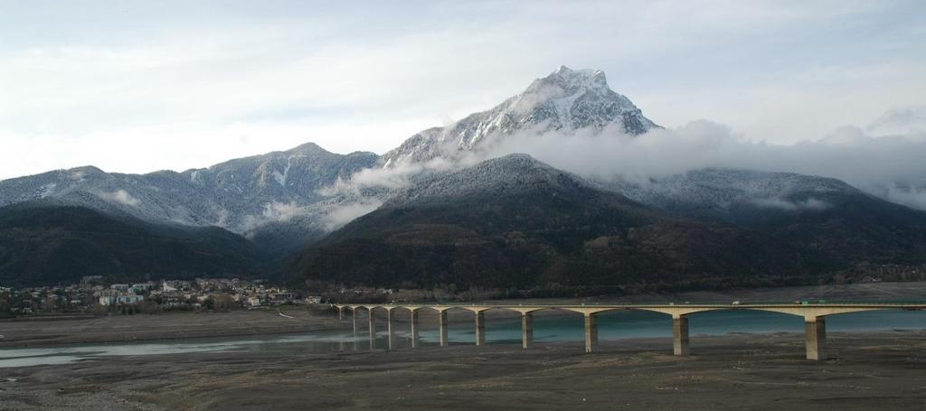 A long bridge crosses a river with snow-covered mountains and a small town in the background.