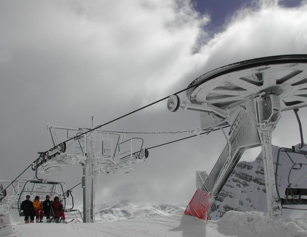 A ski lift covered in snow transports four people up a mountain under a cloudy sky.
