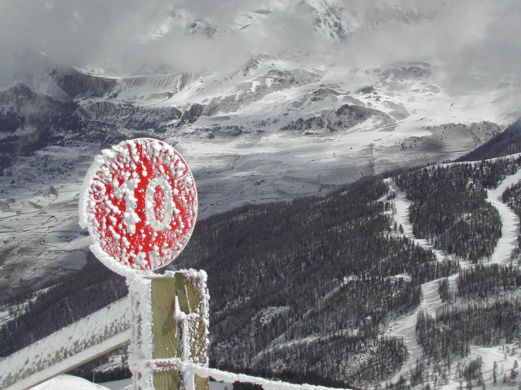 A red speed limit sign covered in snow stands in a snowy mountain landscape with ski trails in the background.