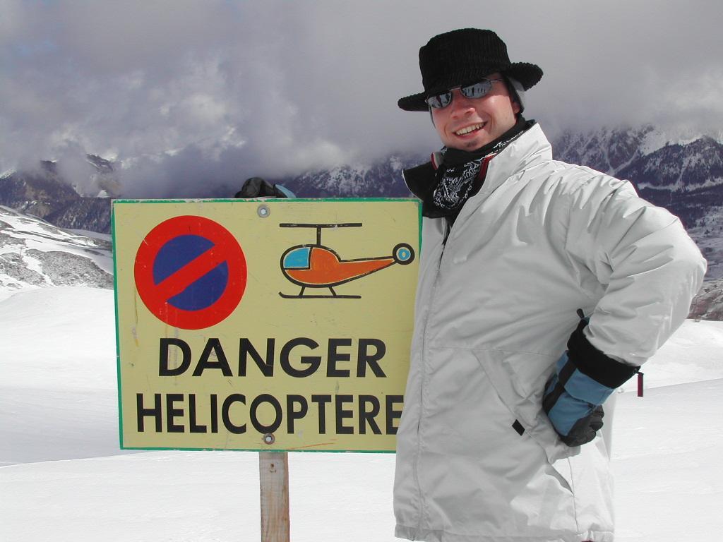 A person in winter clothing smiles next to a "Danger Helicopter" sign in a snowy mountain setting.