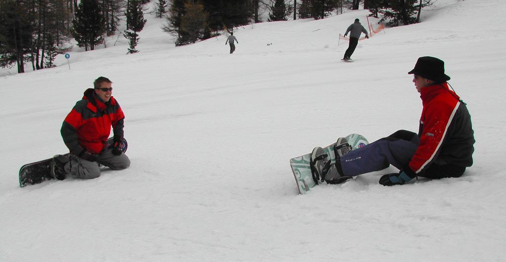 Two snowboarders sit on the snow, facing each other, while another person snowboards downhill in the background.