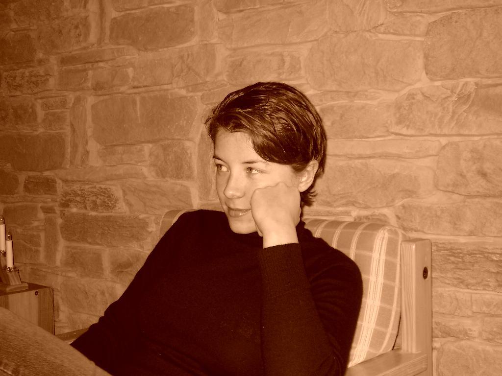 A person in a black sweater sits in a wooden chair, resting their head on their hand and looking away.