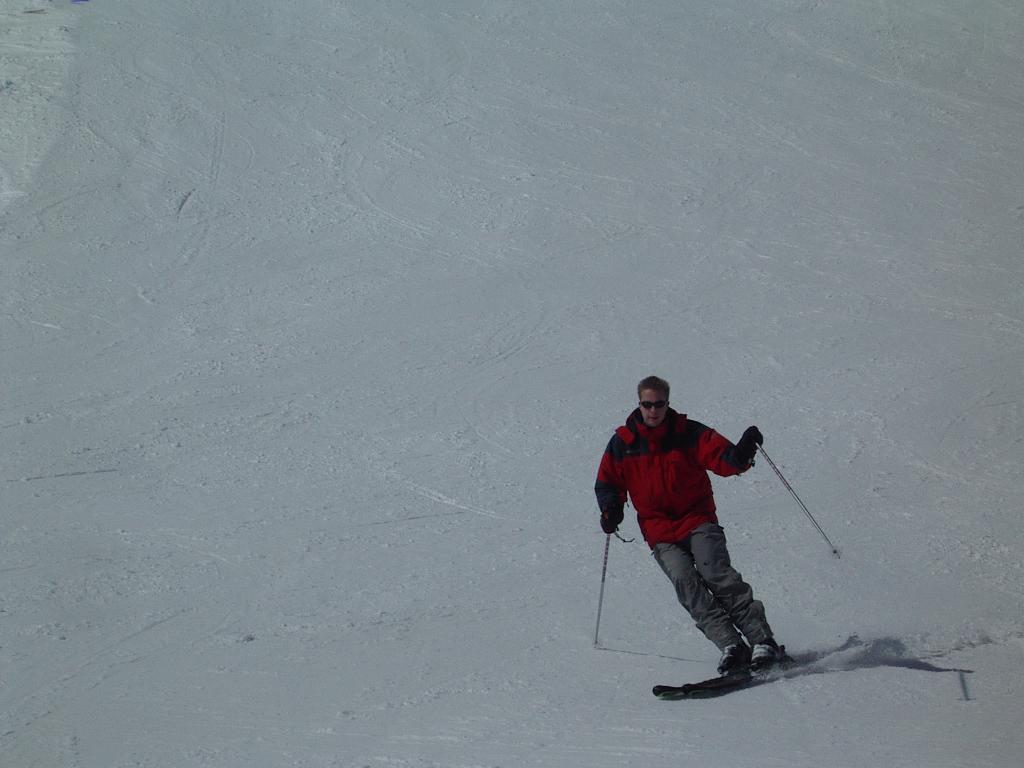 A person wearing a red jacket and sunglasses skis down a snowy slope, using ski poles for balance.