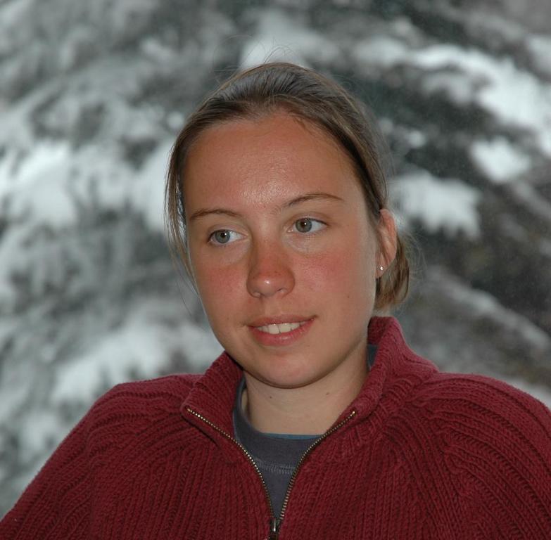 A young woman in a red sweater sits indoors, looking slightly to the side with a neutral expression.