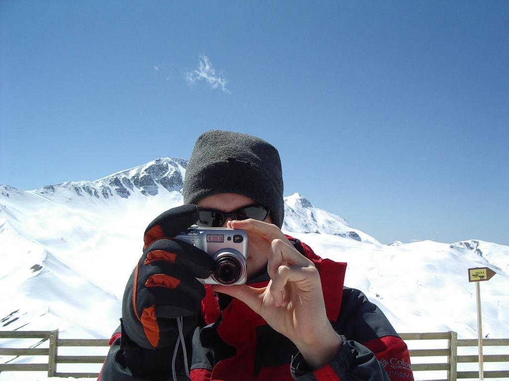 A person in winter clothing holds a camera and takes a photo in a snowy mountain setting.