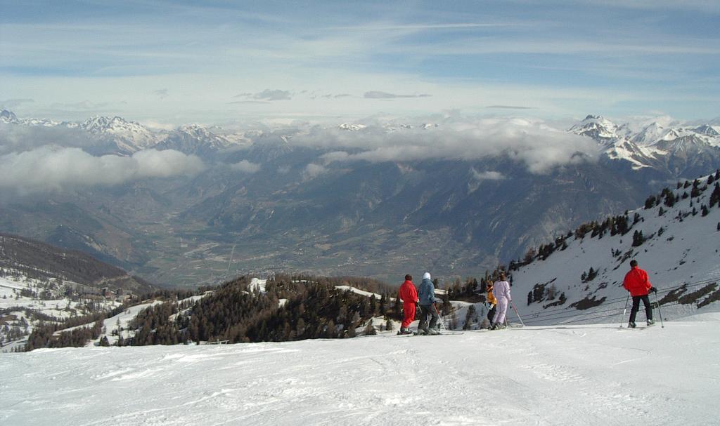 A group of skiers pauses on a snowy slope, looking down at a village in the valley below.
