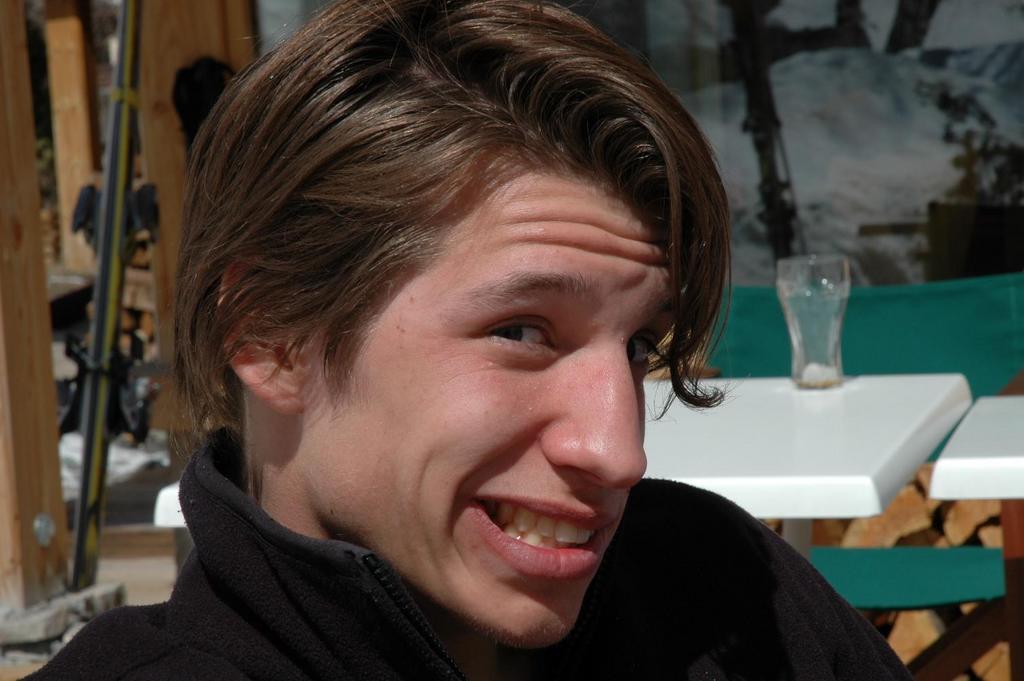 A person with medium-length brown hair smiles while sitting outdoors near tables and chairs.