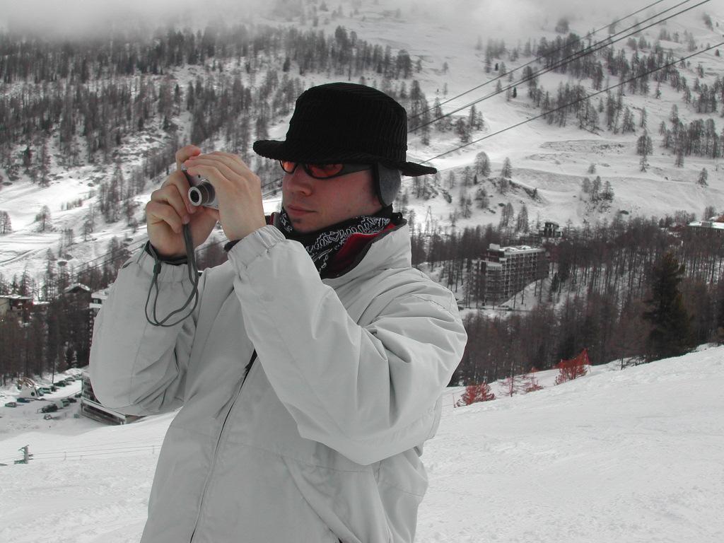A person in a white jacket and black hat takes a photo with a small camera in a snowy landscape.