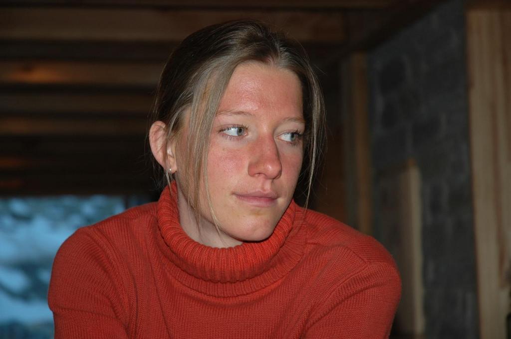 A woman in a red turtleneck sweater looks to the side with a neutral expression indoors.