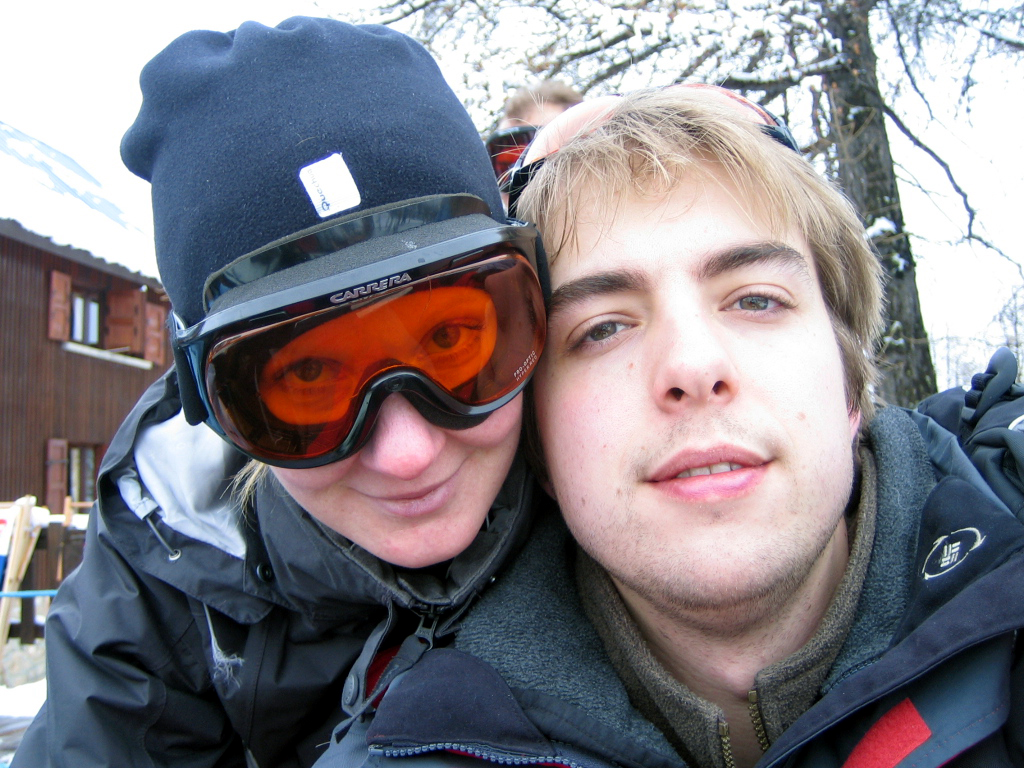 Two people in winter gear take a close-up selfie outdoors, one wearing ski goggles and a beanie.