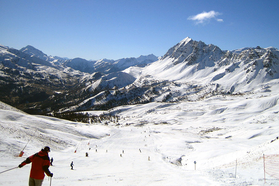 Skiers descend a snowy mountain slope with a clear blue sky and rugged peaks in the background.