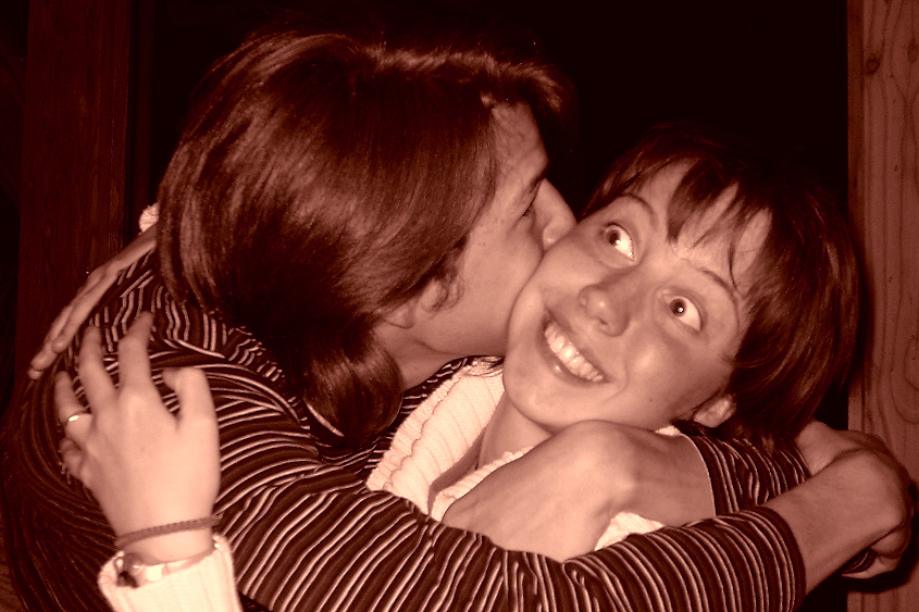 One person hugs and kisses another on the cheek, while the other smiles and looks away playfully.