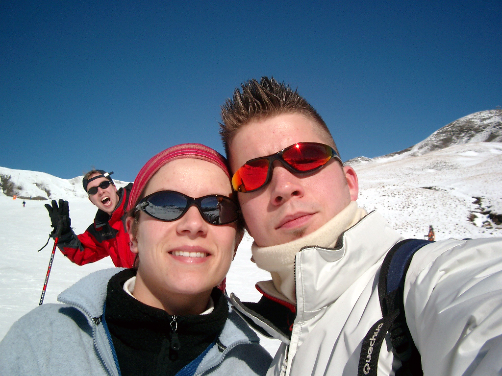 Two people in sunglasses take a close-up selfie on a snowy mountain, while another person playfully poses behind them.