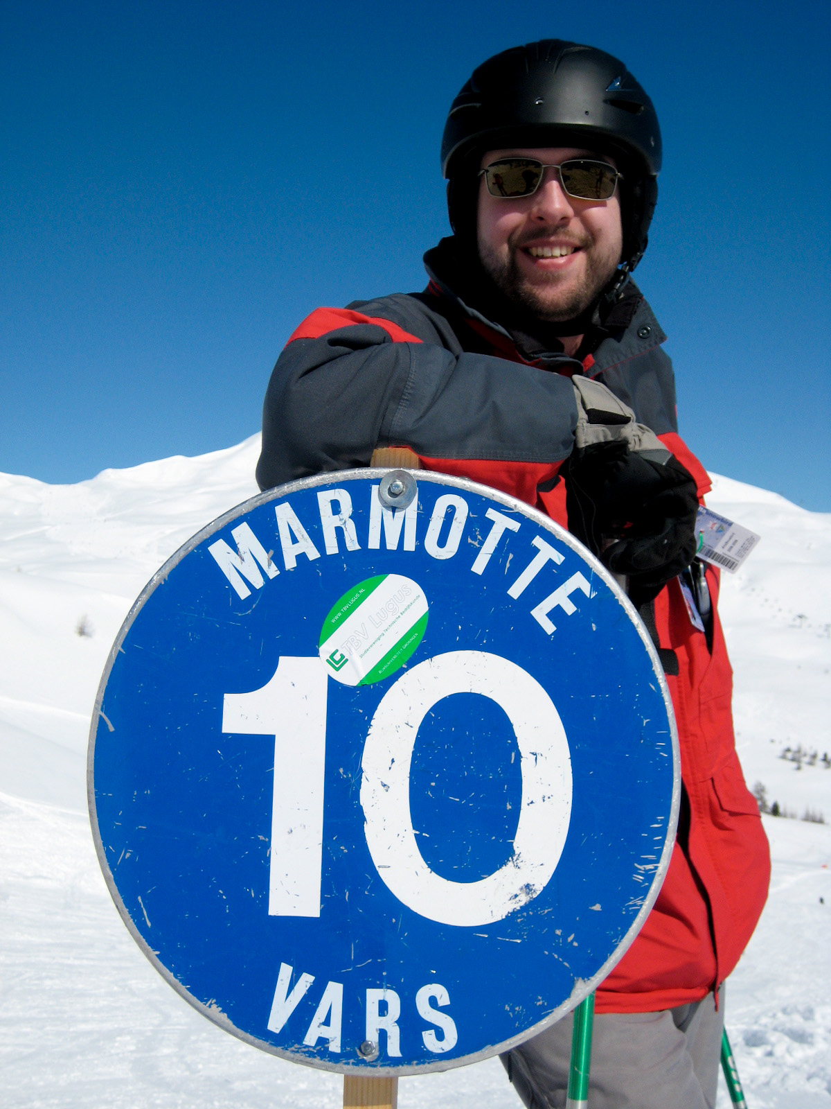 A person in ski gear smiles while leaning on a blue "Marmotte 10 Vars" sign on a snowy slope.