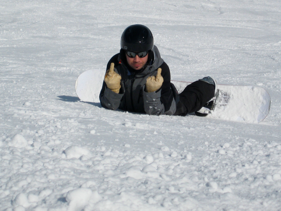 A snowboarder in a helmet and goggles lies on the snow, raising both hands with middle fingers extended.