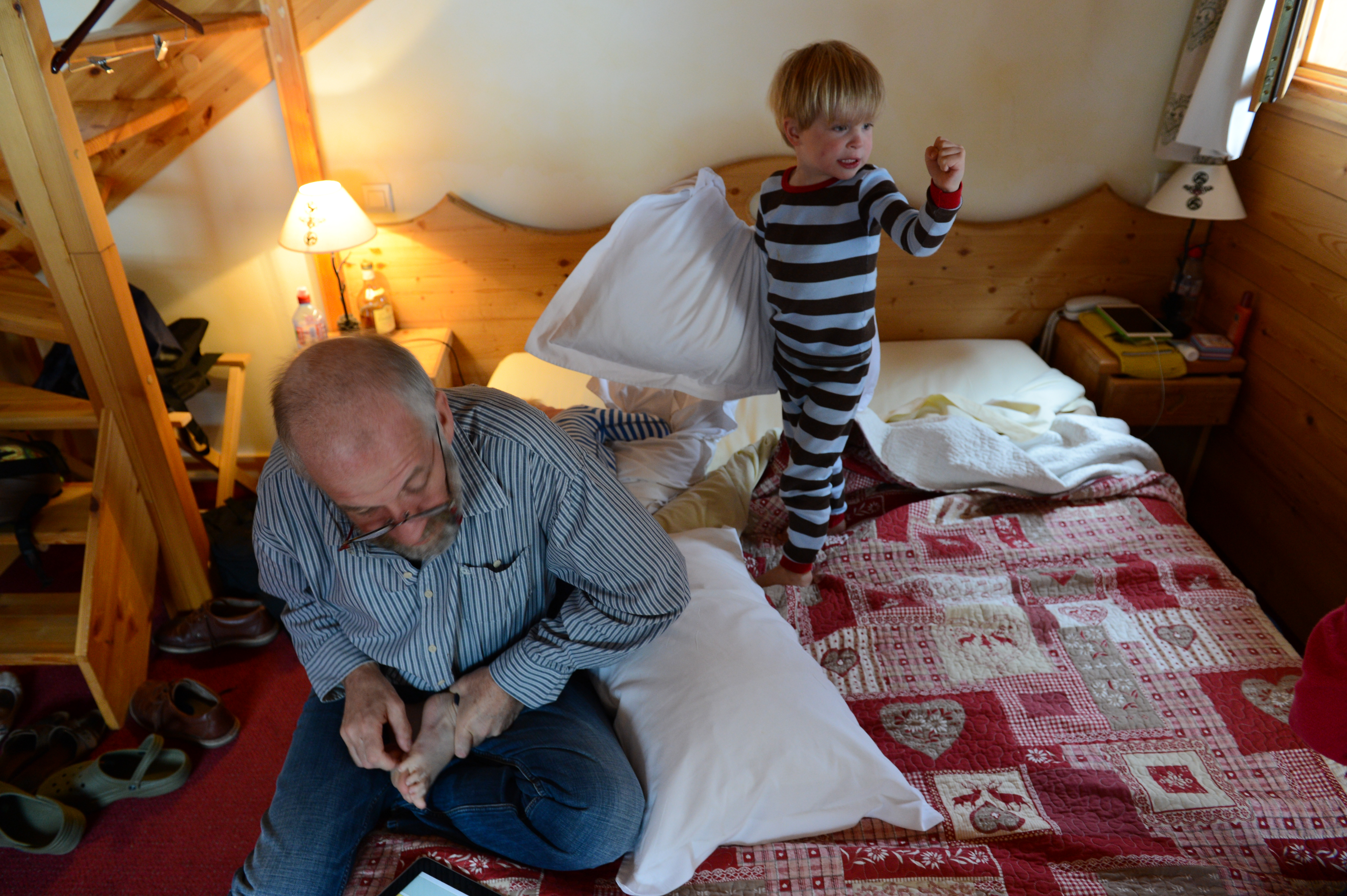 A child in striped pajamas holds a pillow, appearing ready to play, while an adult sits on the bed.