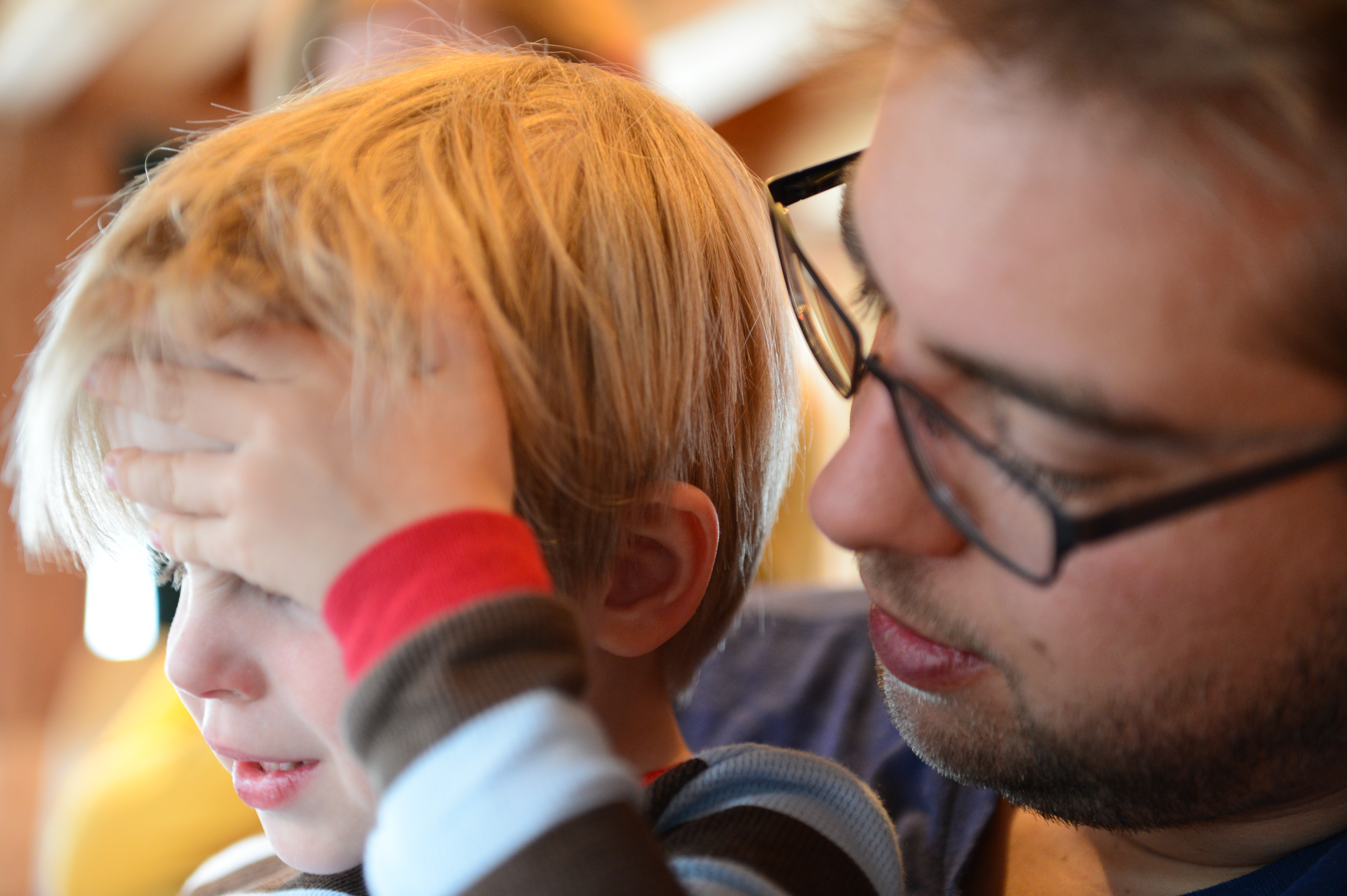 A child covers their face while an adult wearing glasses looks at them closely.