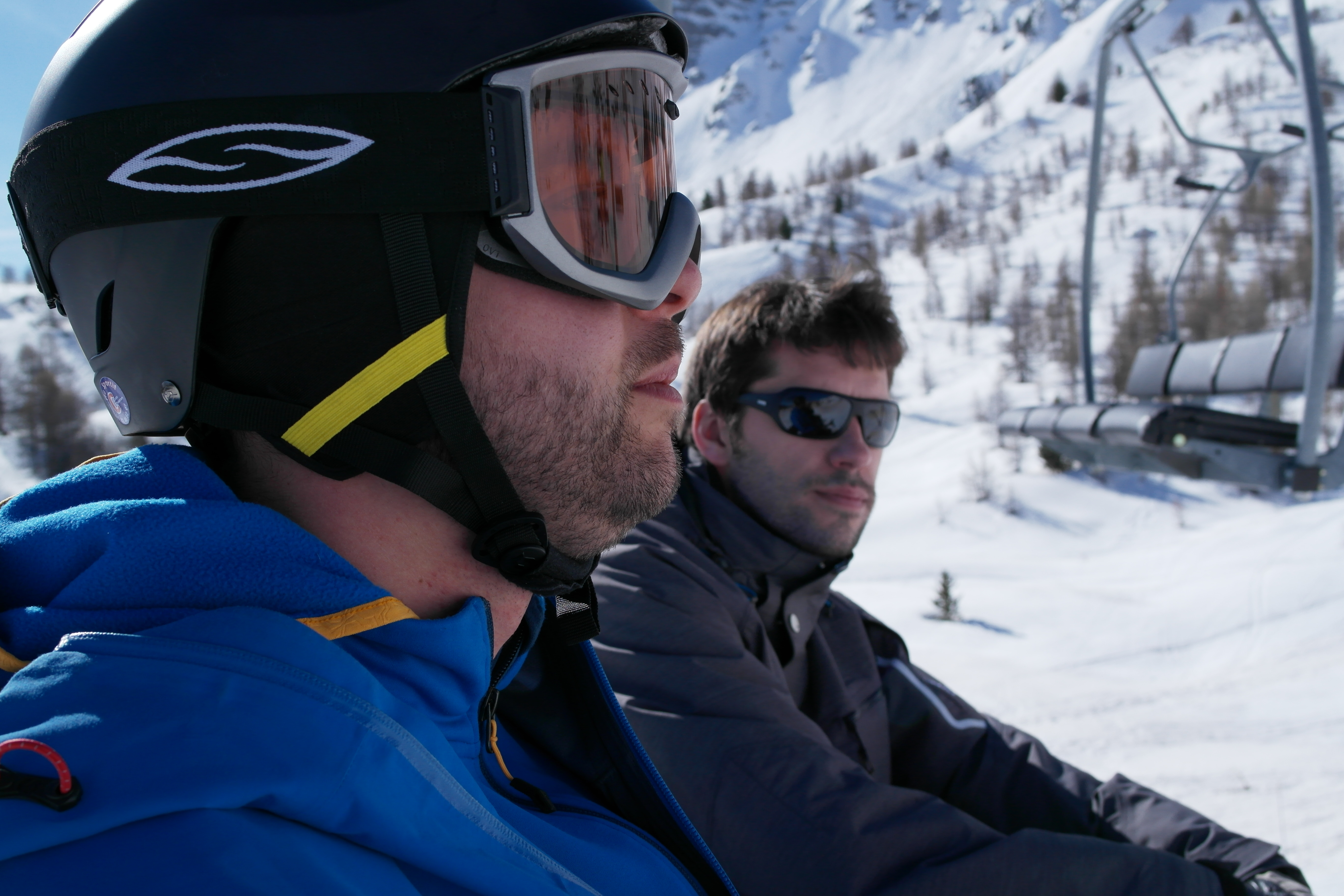 Two men wearing winter gear and sunglasses ride a ski lift in a snowy mountain setting.
