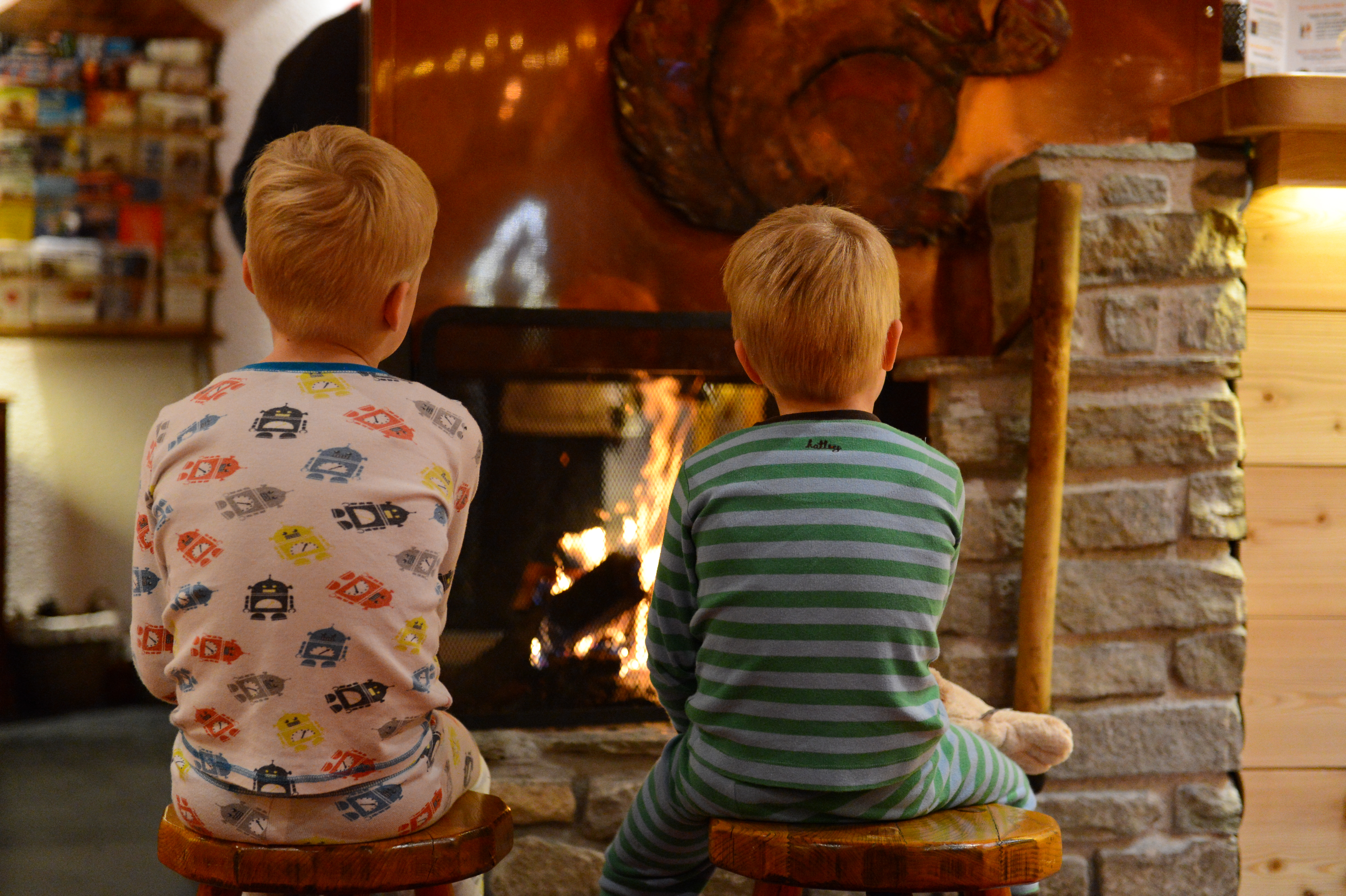 Two children in pajamas sit on stools, facing a warm fireplace in a cozy indoor setting.
