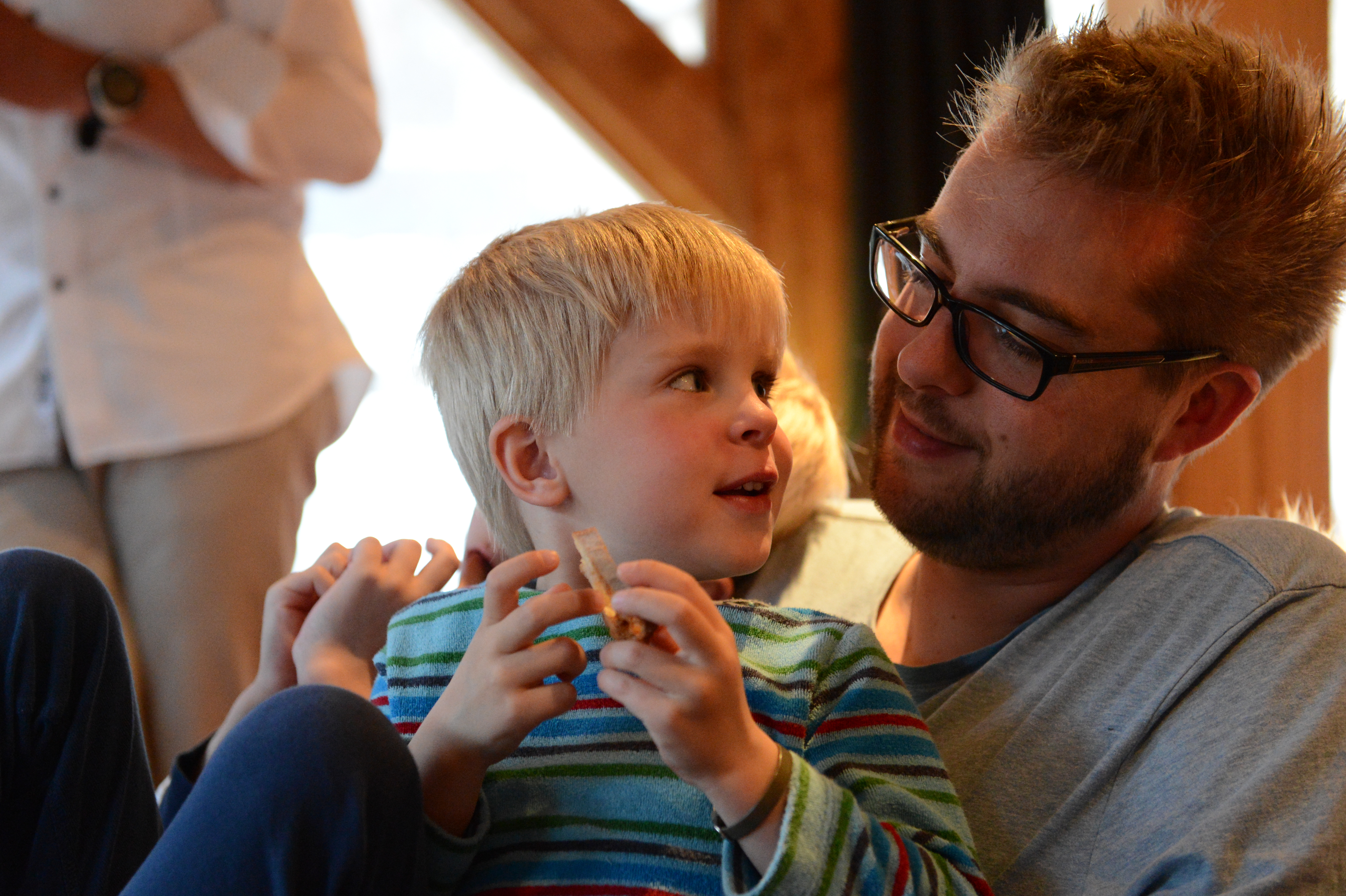 A man with glasses smiles while holding a young child in a striped shirt, who is eating a snack.