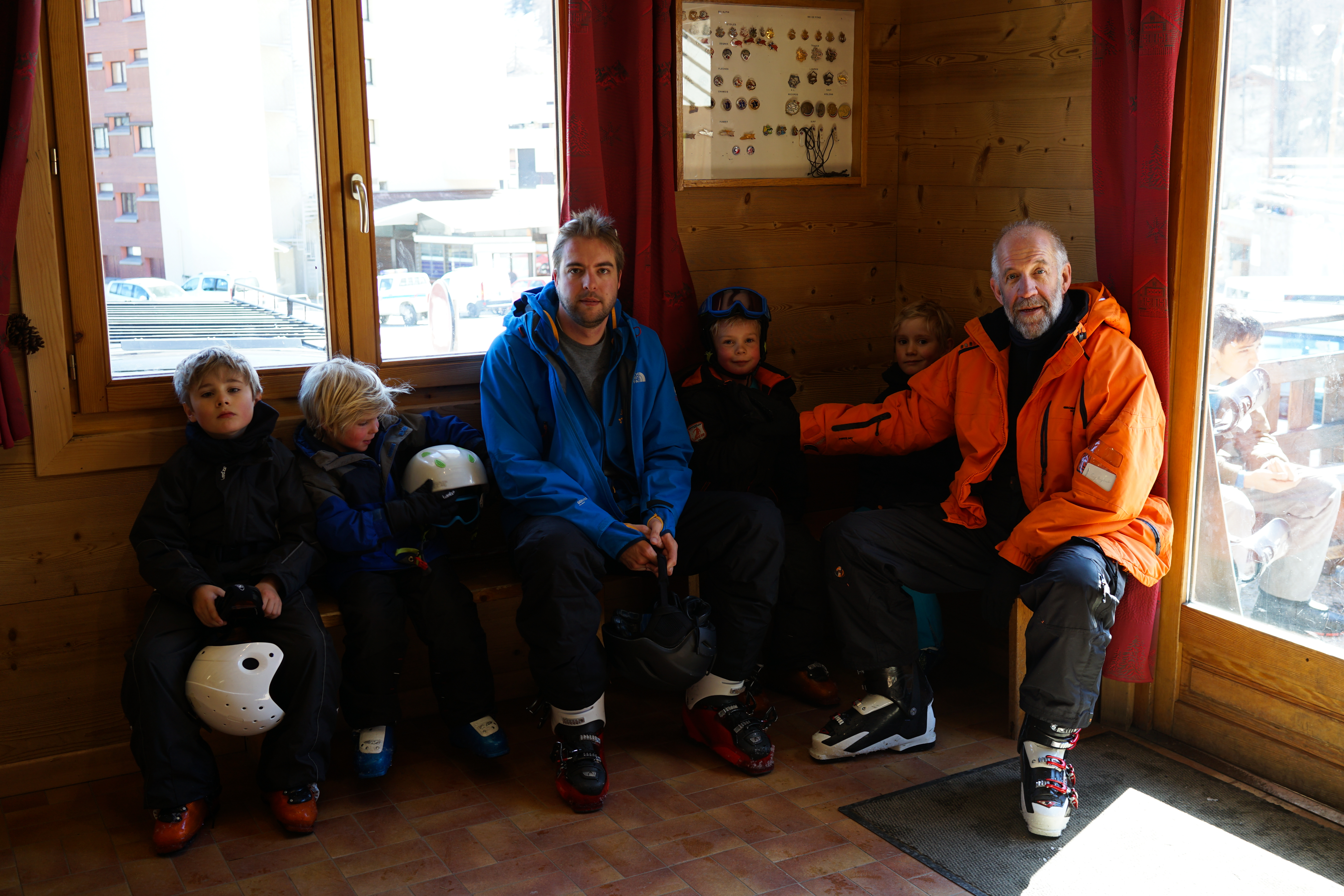 A group of adults and children in ski gear sit on a bench inside a wooden cabin, holding helmets.