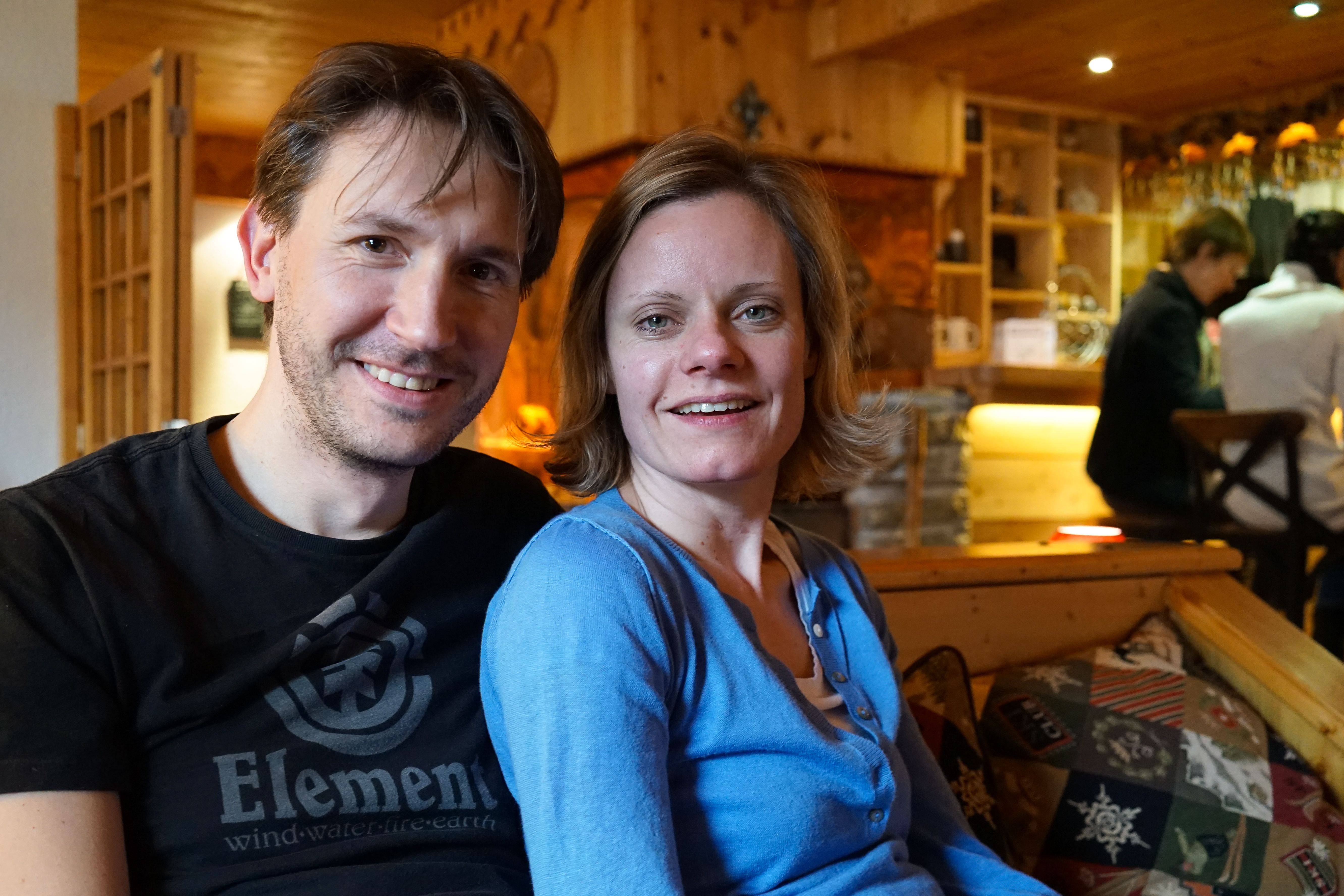 A man and a woman sit closely together on a couch, smiling at the camera in a cozy setting.