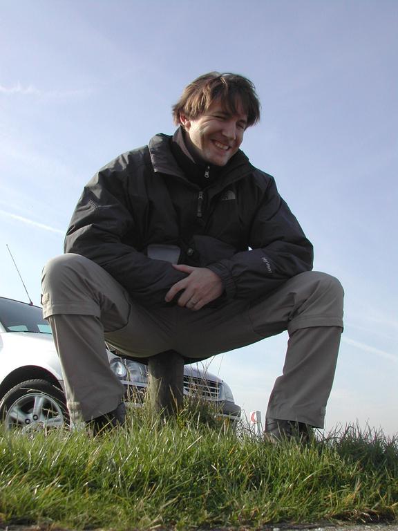A man in a jacket squats on grass, smiling.
