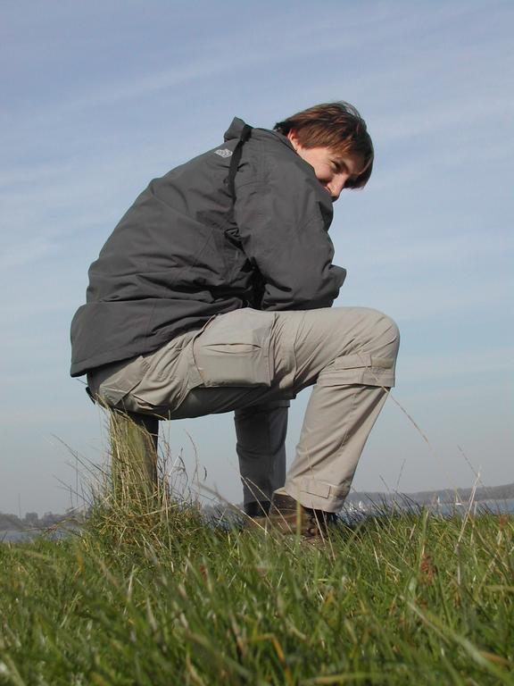 A person in a dark jacket and beige pants sits on a wooden post, looking back over their shoulder.