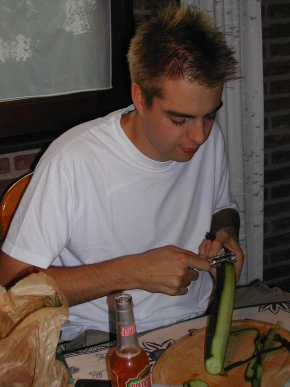 A person in a white shirt is peeling a cucumber at a table with a cutting board and bottle.