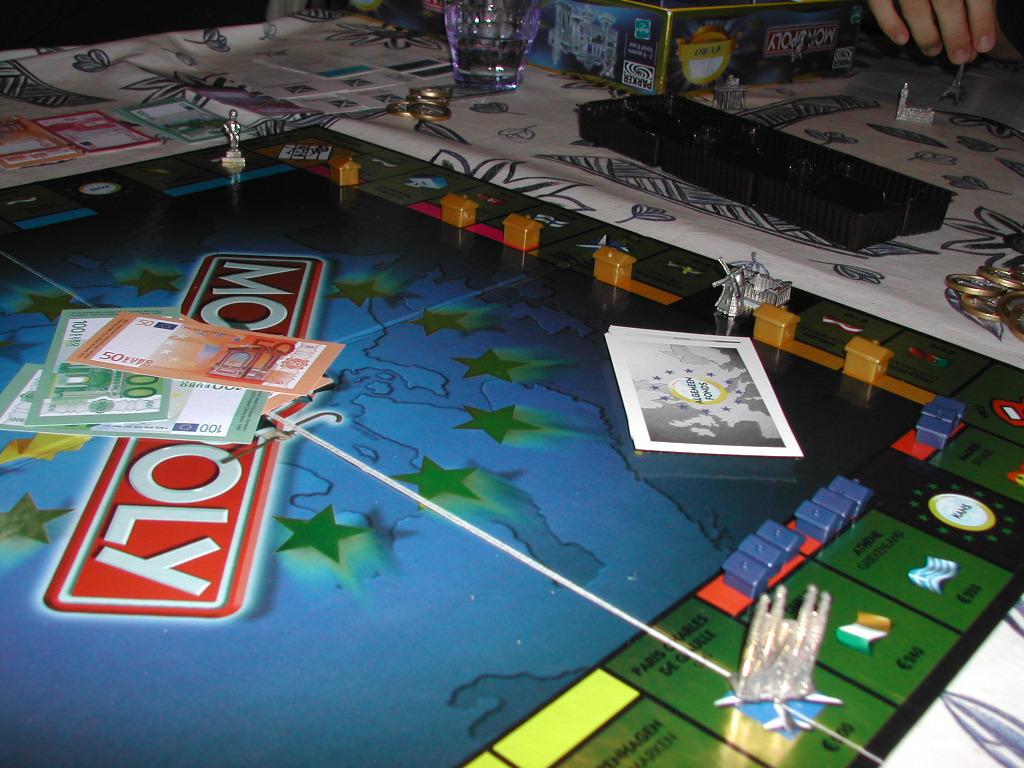 A Monopoly board game with Euro-themed currency, metal playing pieces, and cards on a table with players' hands visible.