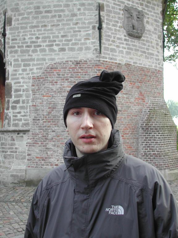 A person wearing a black jacket and hat stands in front of an old brick building with a crest.