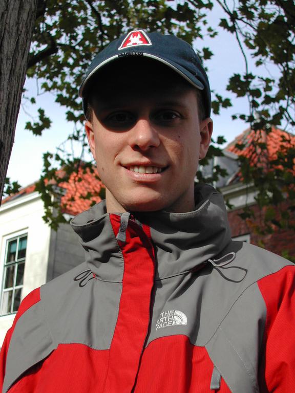 A person wearing a red and gray jacket and a cap smiles at the camera outdoors with trees and buildings behind.