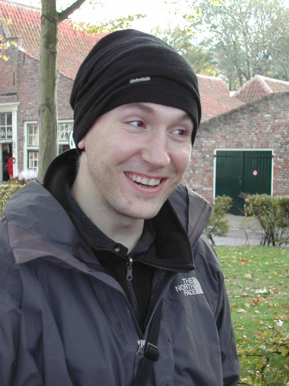 A person wearing a black beanie and jacket smiles while standing outdoors near brick buildings and greenery.