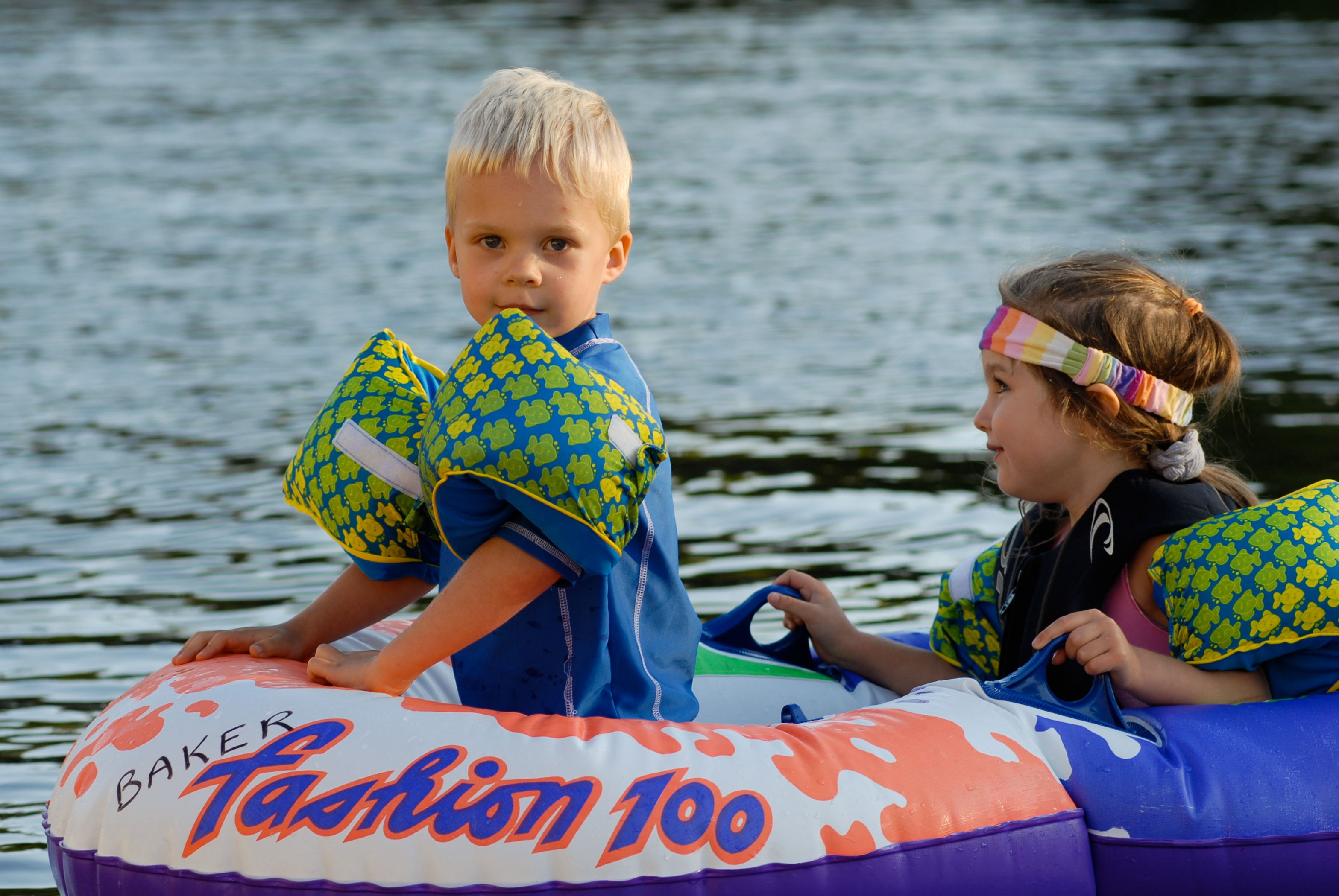 Two young children wearing floatation gear sit in an inflatable boat on the water, holding onto the handles.