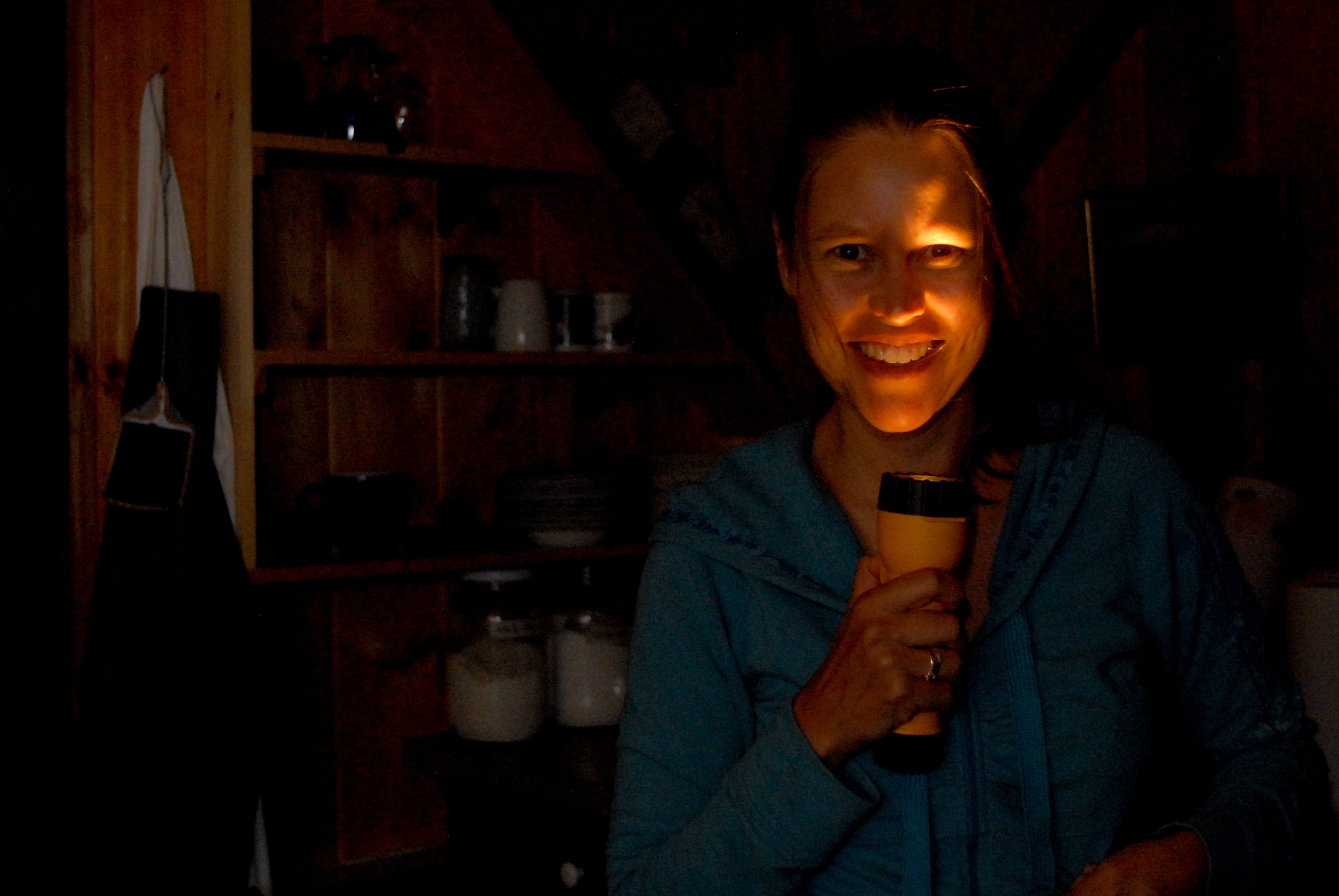 A woman in a dark room holds a flashlight under her face, creating a spooky lighting effect.