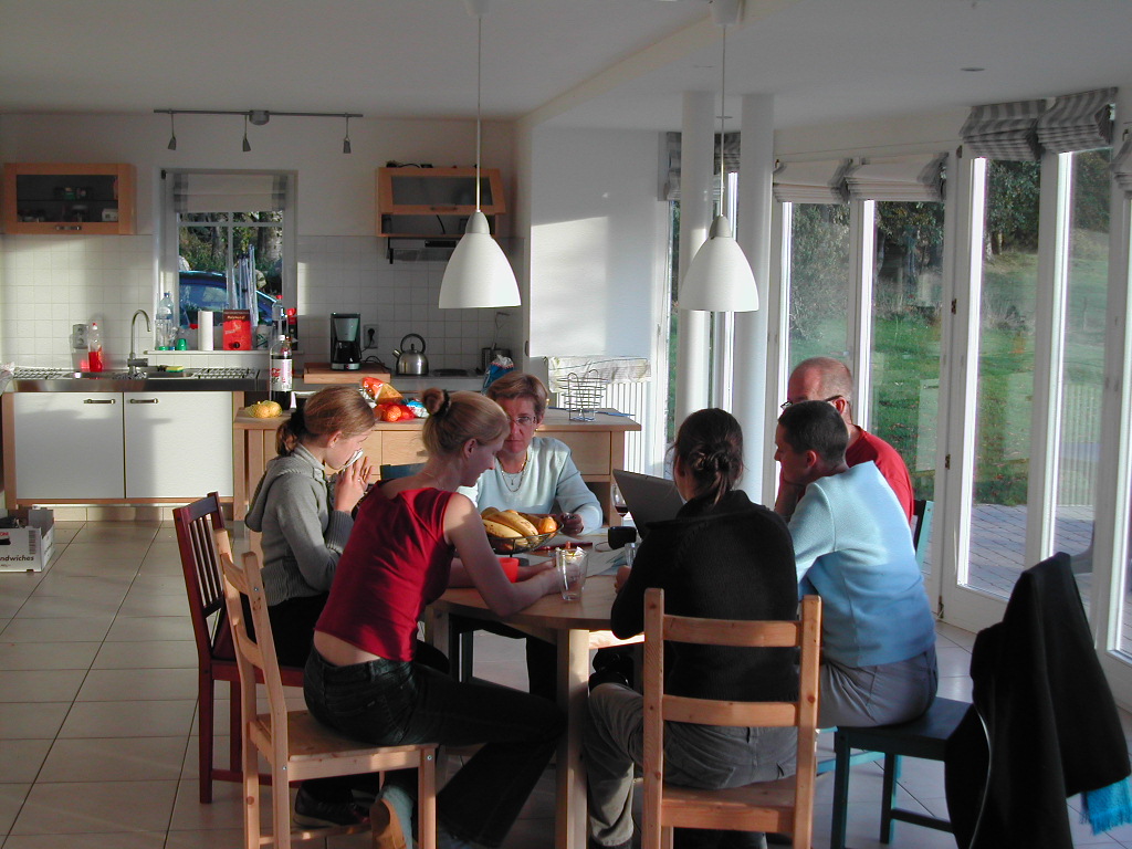 Six people sit around a wooden table in a bright kitchen, eating and talking.