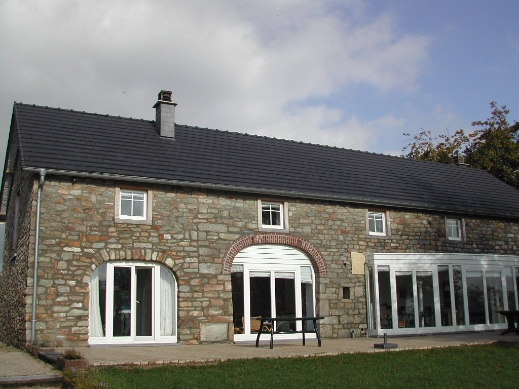 A stone house with a dark roof, large windows, and a patio with tables in front.