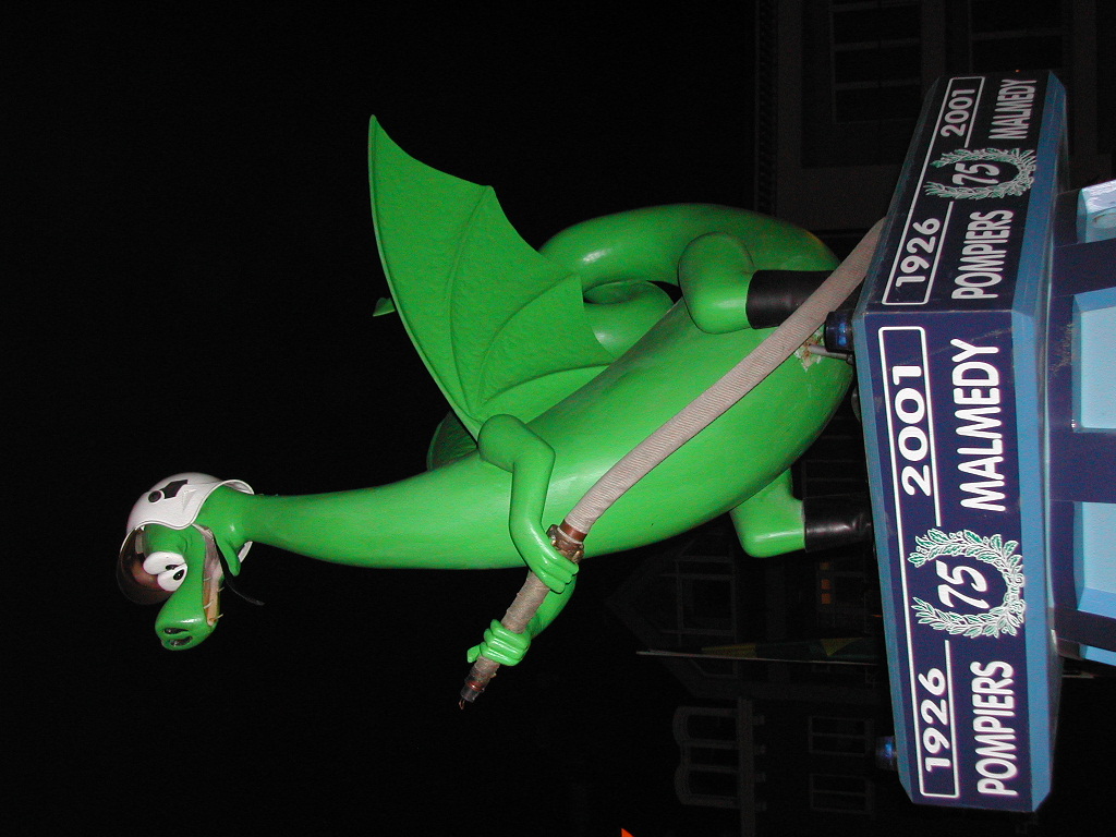 A green cartoon dragon wearing a helmet holds a fire hose while attached to a decorated structure at night.