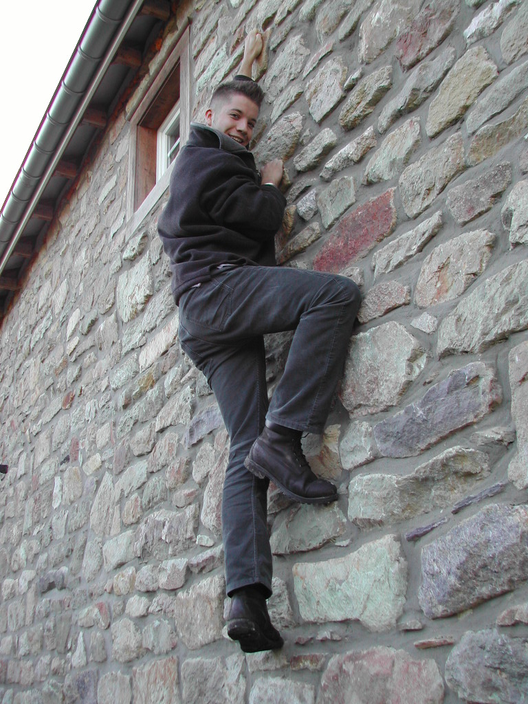A person in dark clothing climbs a stone wall, gripping the rocks and looking toward the camera with a smile.