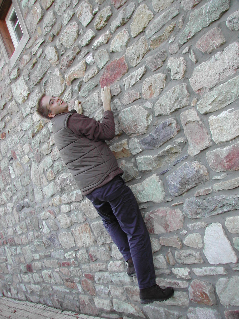 A man wearing glasses and a vest playfully pretends to climb a stone wall while smiling at the camera.