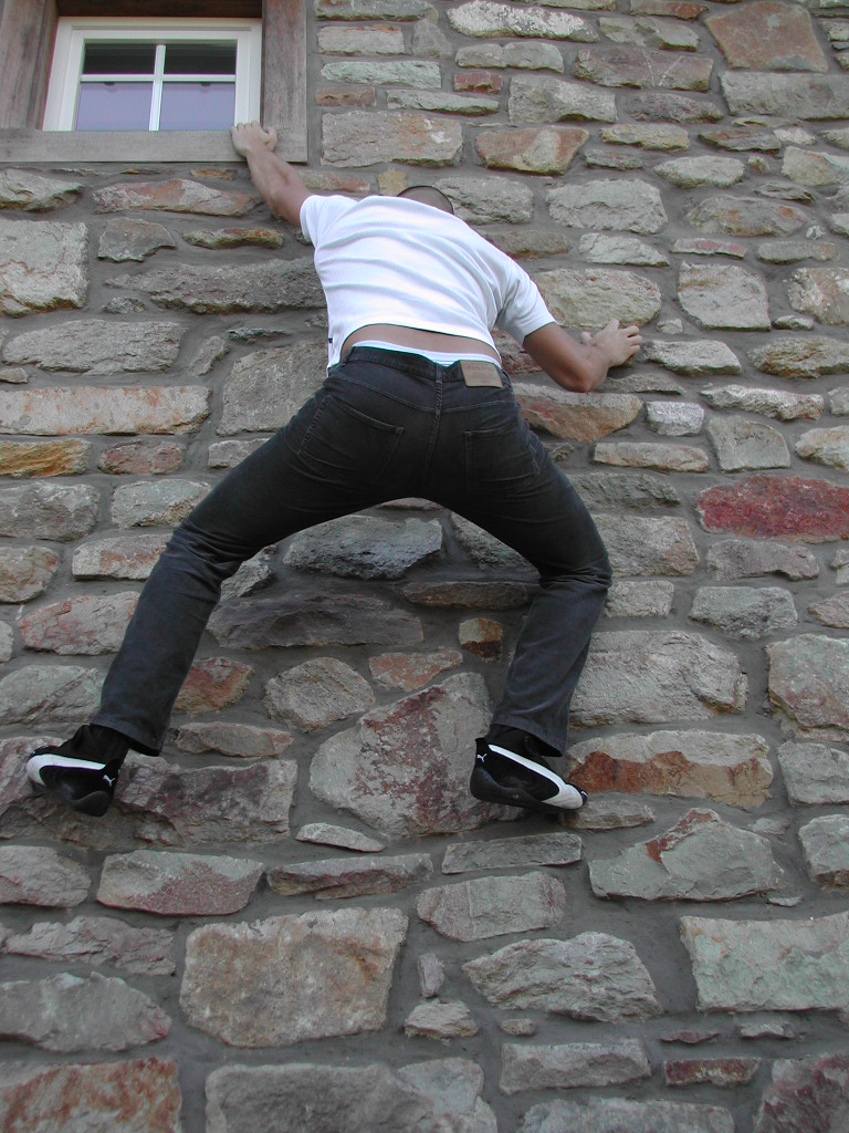 A person in a white shirt and dark pants climbs a stone wall, reaching toward a window above.
